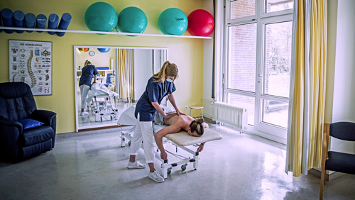 Im Mittelpunkt des Bildes befindet sich eine Behandlungsbank im Therapiezentrum Nord des Klinikums Nordfriesland, an der eine Physiotherapeutin einen Patienten behandelt. 