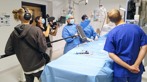 Das Bild zeigt ein Filmteam in dem Angiographieraum der Klinik Husum. Im Vordergrund befinden sich der Tontechniker, dahinter der Kameramann, der eine Szene am Behandlungstisch des Angiographiegerätes filmt. Zwei in OP-Kleidung mit Mundschutz und Kopfhaube befindliche Ärzte erläutern einen medizinischen Sachverhalt.