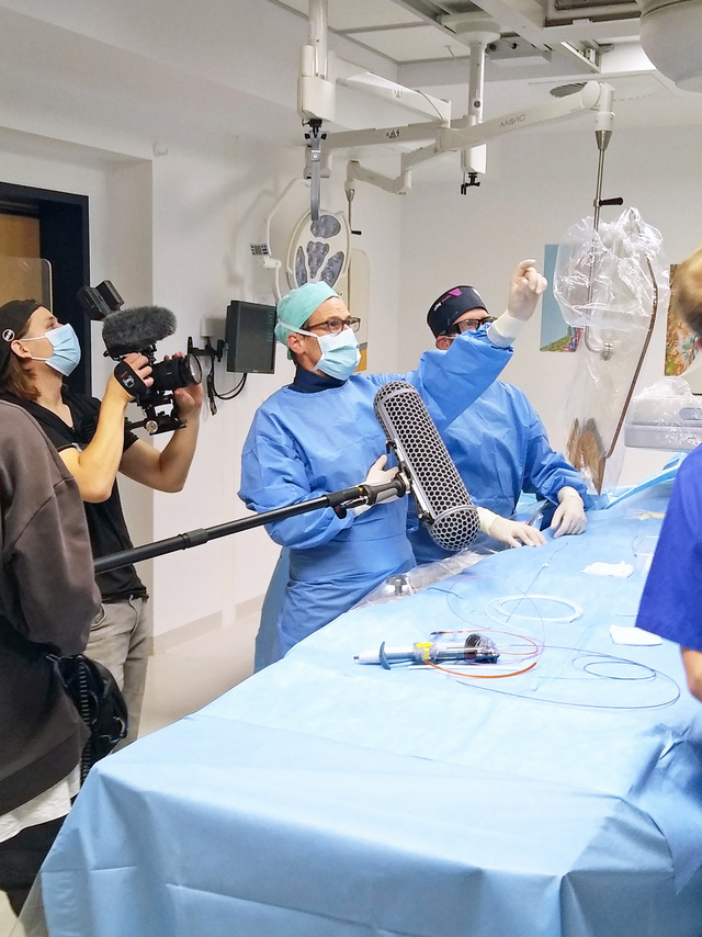 Das Bild zeigt ein Filmteam in dem Angiographieraum der Klinik Husum. Im Vordergrund befinden sich der Tontechniker, dahinter der Kameramann, der eine Szene am Behandlungstisch des Angiographiegerätes filmt. Zwei in OP-Kleidung mit Mundschutz und Kopfhaube befindliche Ärzte erläutern einen medizinischen Sachverhalt.